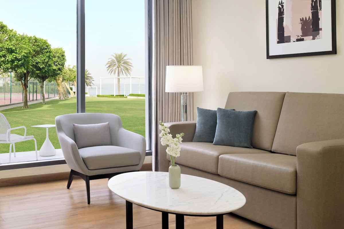 A modern living room with a beige sofa, a grey armchair, and a white coffee table. A vase with white flowers is on the table. A large window offers a view of a green lawn and palm trees.