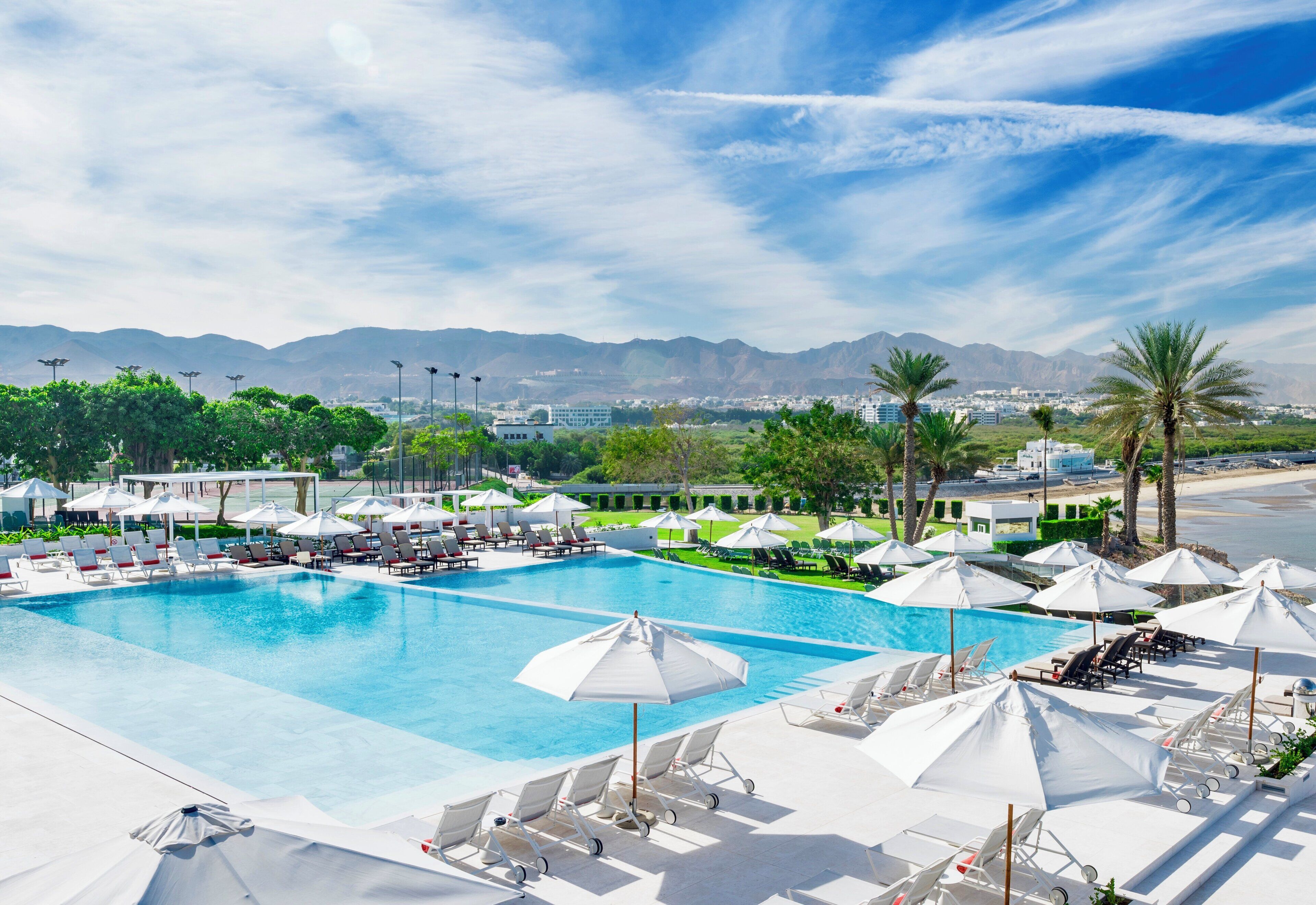 A large swimming pool with sun loungers and umbrellas, surrounded by palm trees and mountains in the background.