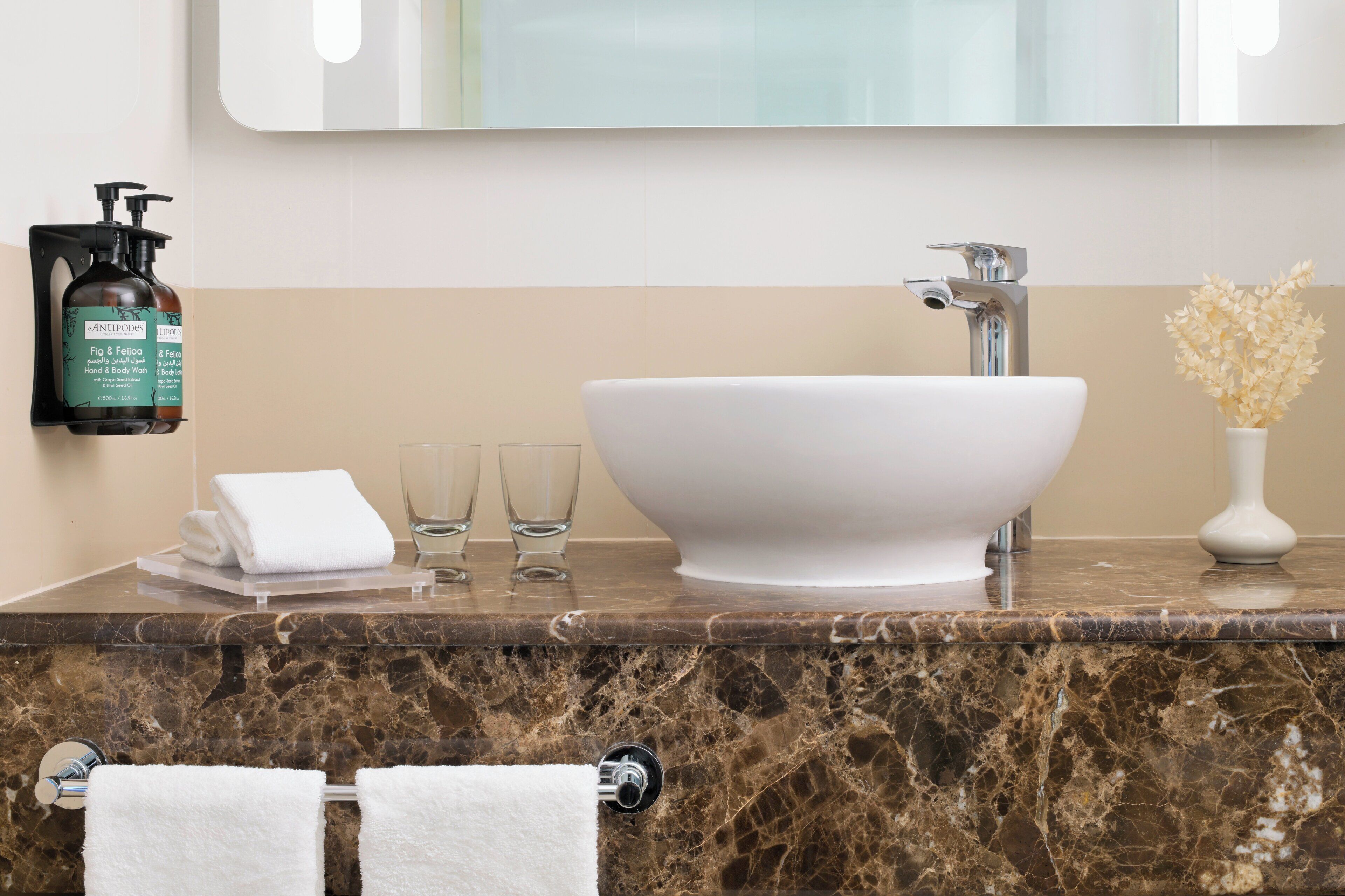A bathroom with a marble countertop, a white sink, a mirror, a towel rack, a glass of water, and a bottle of hand soap.