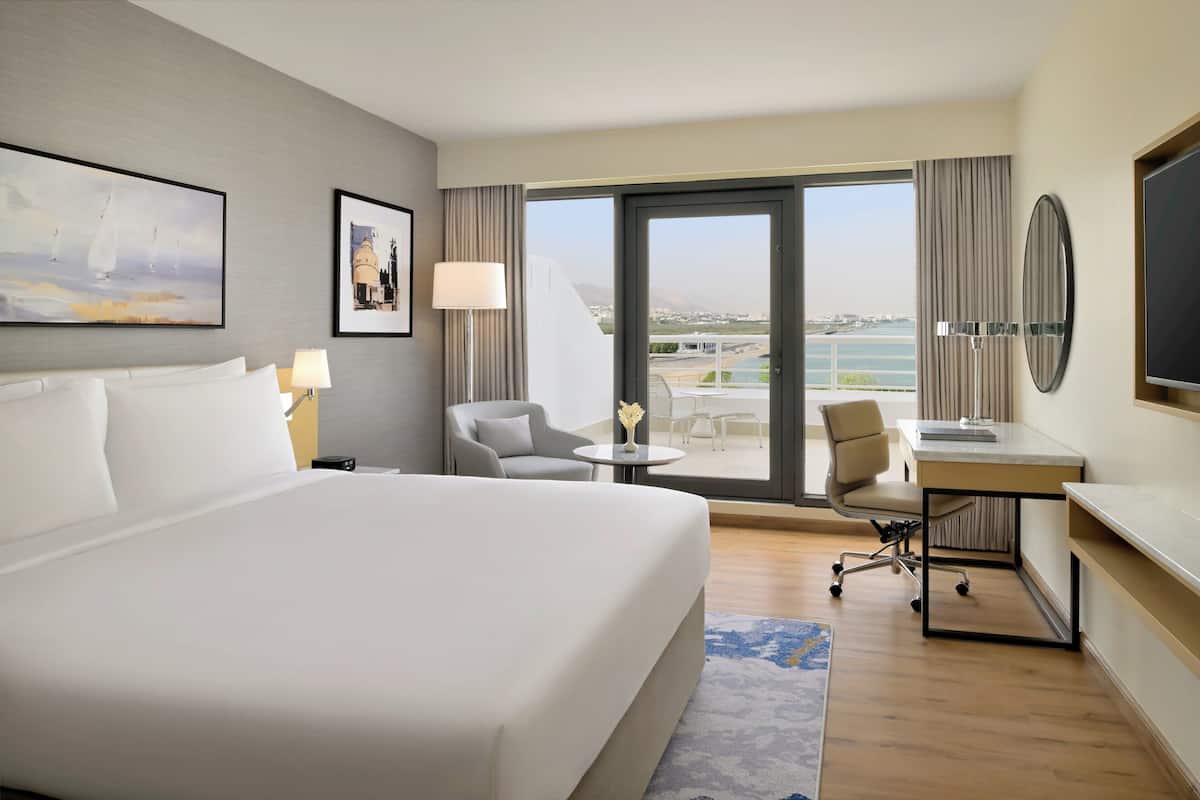 A hotel room with a large bed, a desk, a chair, a small table, and a view of the city and water.