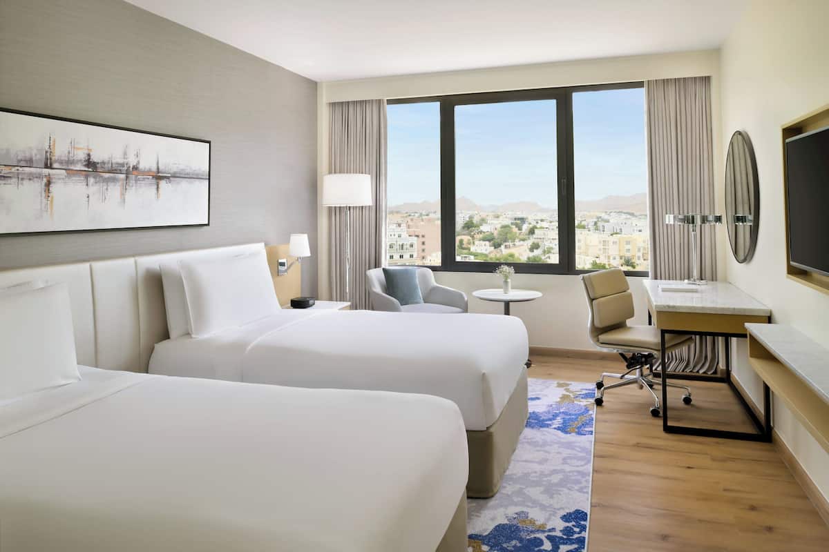 A hotel room with two beds, a desk, a chair, a TV, and a large window with a city view.