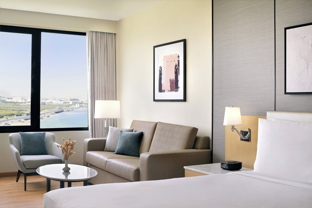 A hotel room with a bed, two armchairs, a small table with a vase, a nightstand with a lamp, and a large window offering a city view.