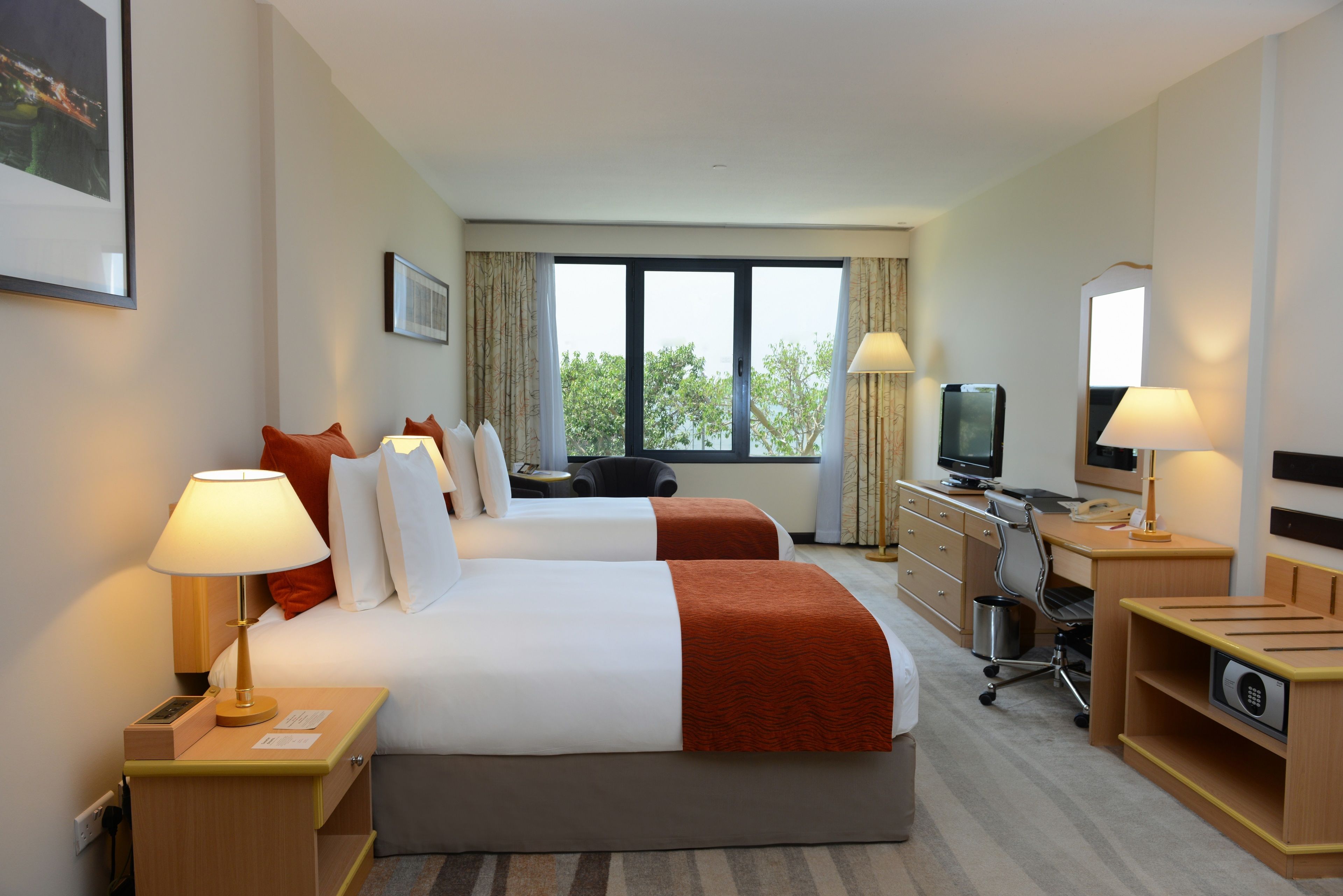 A hotel room with two beds, a desk with a computer, a TV, and a window with a view of trees.