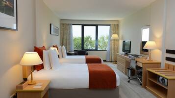 A hotel room with two beds, a desk with a computer, a TV, and a window with a view of trees.