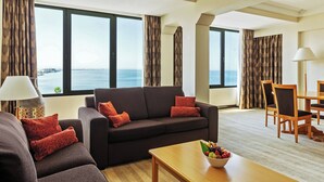 A modern living room with a sofa, coffee table, and dining area. Large windows offer a view of the sea.