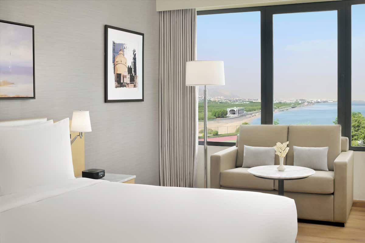 A hotel room with a bed, a sofa, a table with a vase, a lamp, and a view of the sea and buildings.
