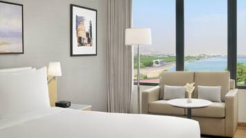 A hotel room with a bed, a sofa, a table with a vase, a lamp, and a view of the sea and buildings.
