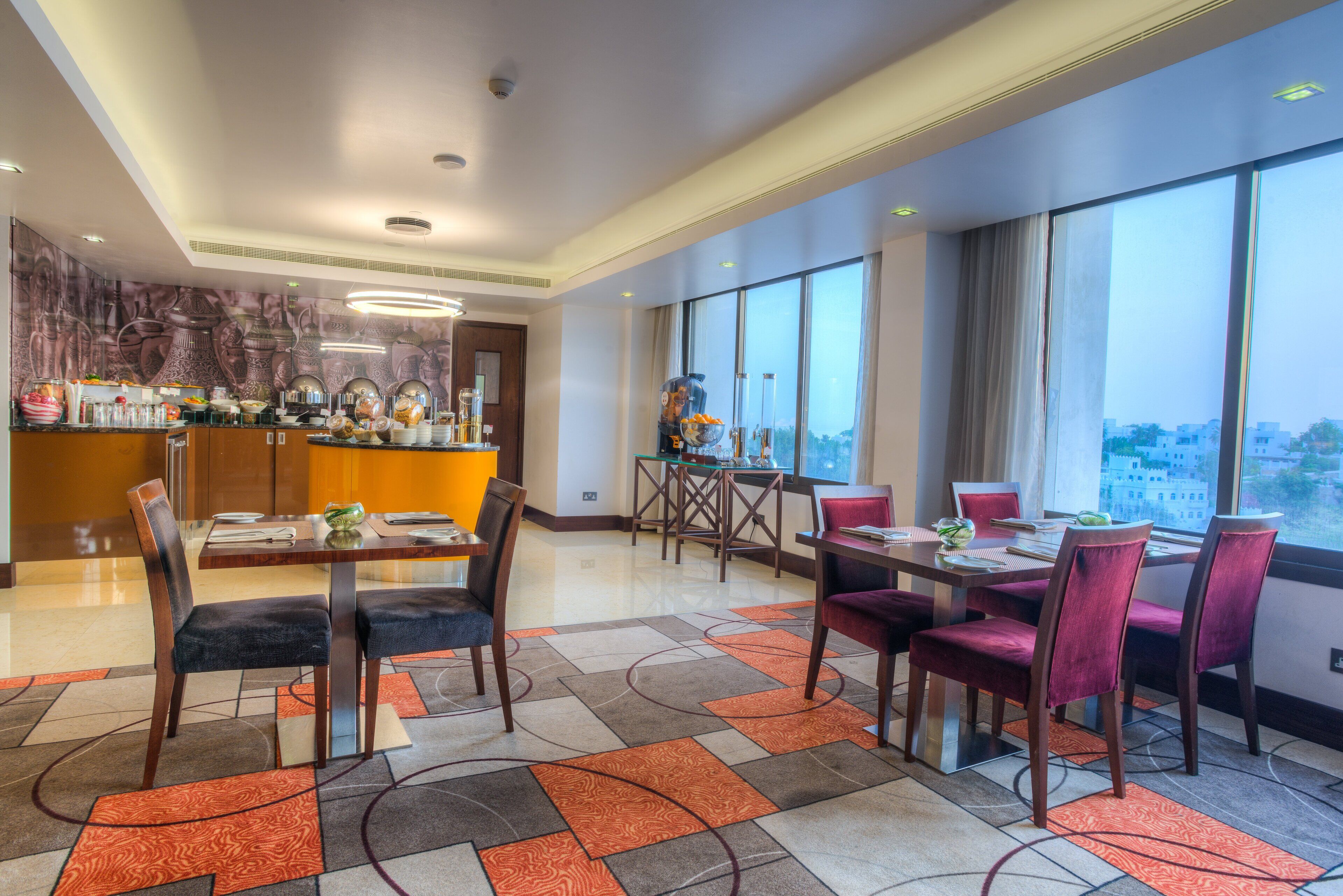 A modern hotel dining area with a large window view, a central bar, and a mix of seating arrangements.