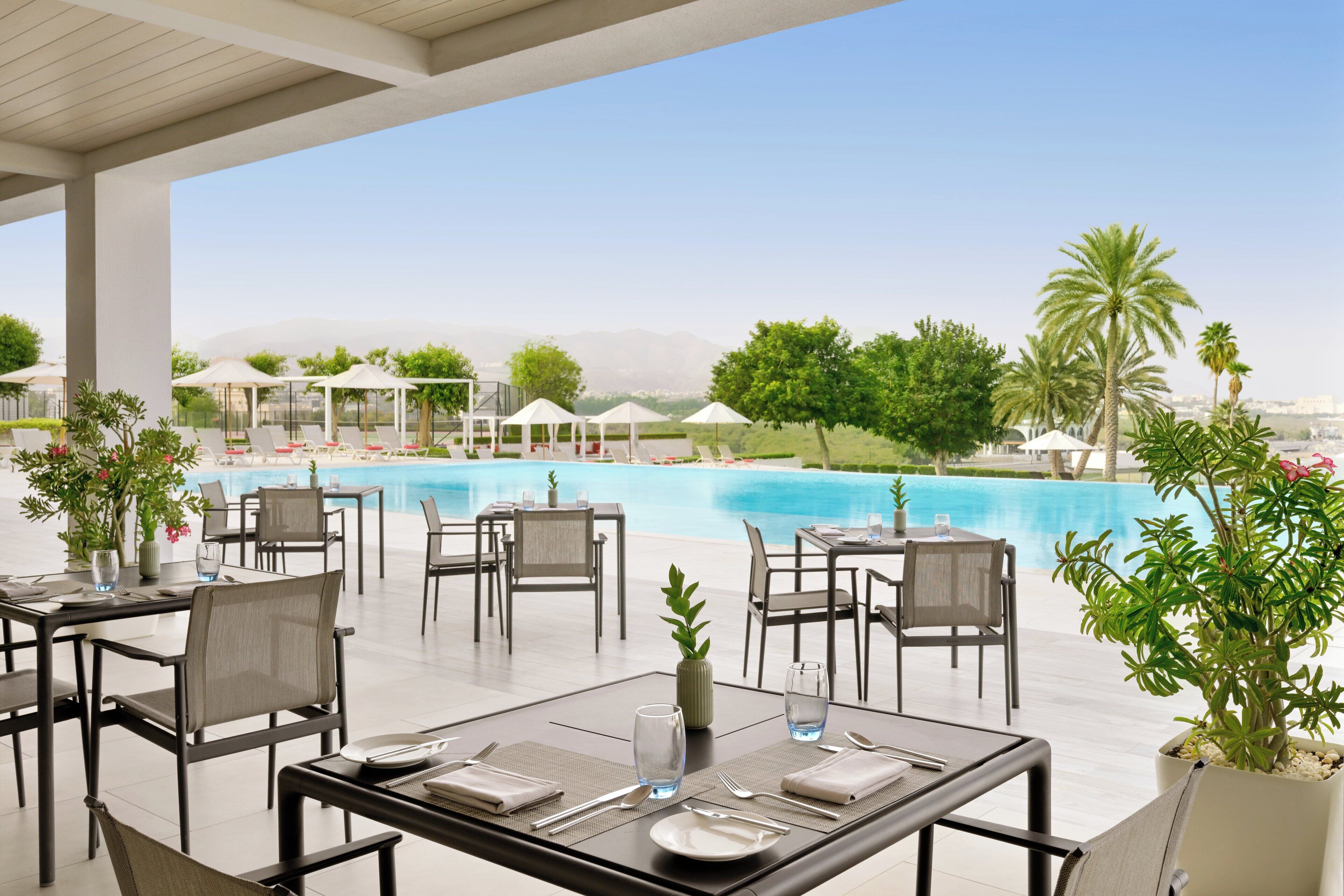 A poolside restaurant with tables and chairs, a pool, and palm trees.