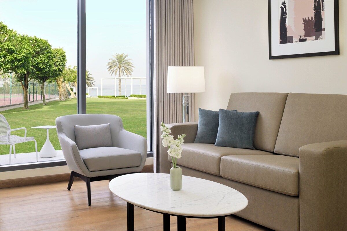 A modern living room with a beige sofa, a grey armchair, and a white coffee table. A vase with white flowers is on the table. A large window offers a view of a green lawn and palm trees.