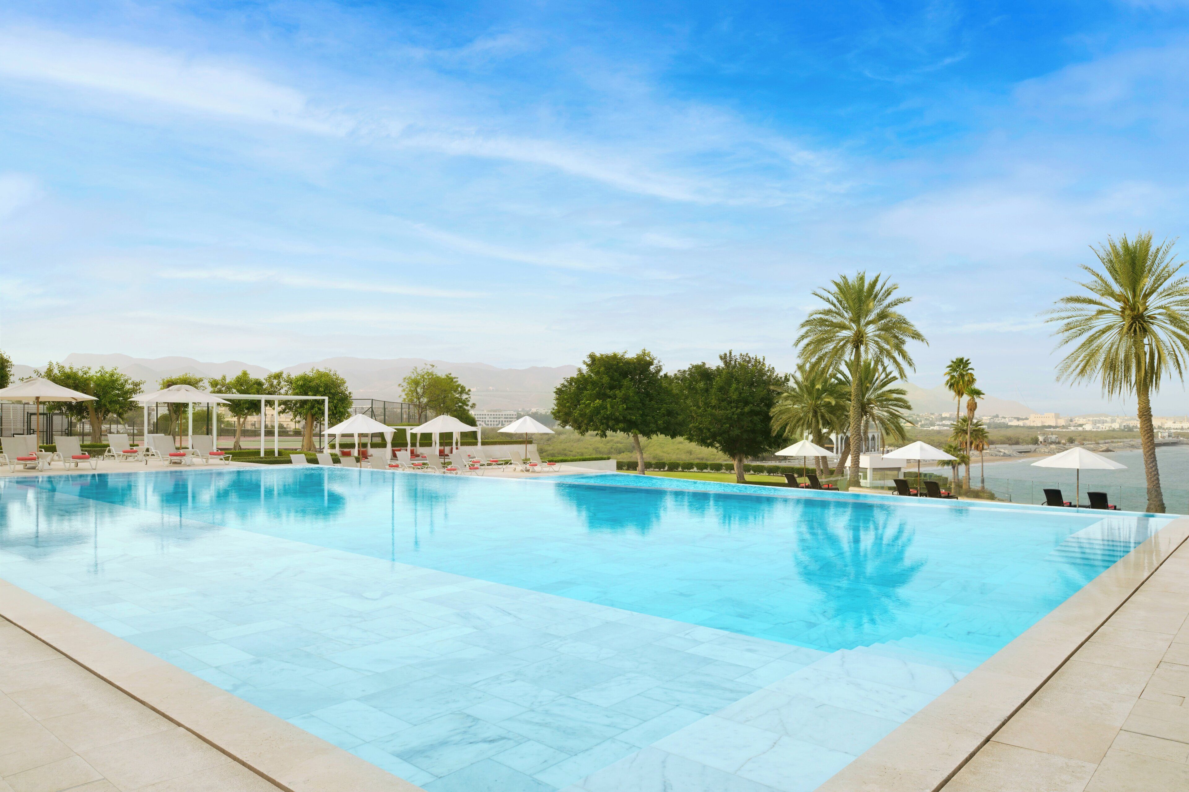 A swimming pool with lounge chairs and umbrellas, surrounded by palm trees and a clear sky.