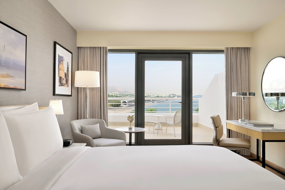 A hotel room with a bed, two chairs, a desk, a lamp, and a view of the sea.