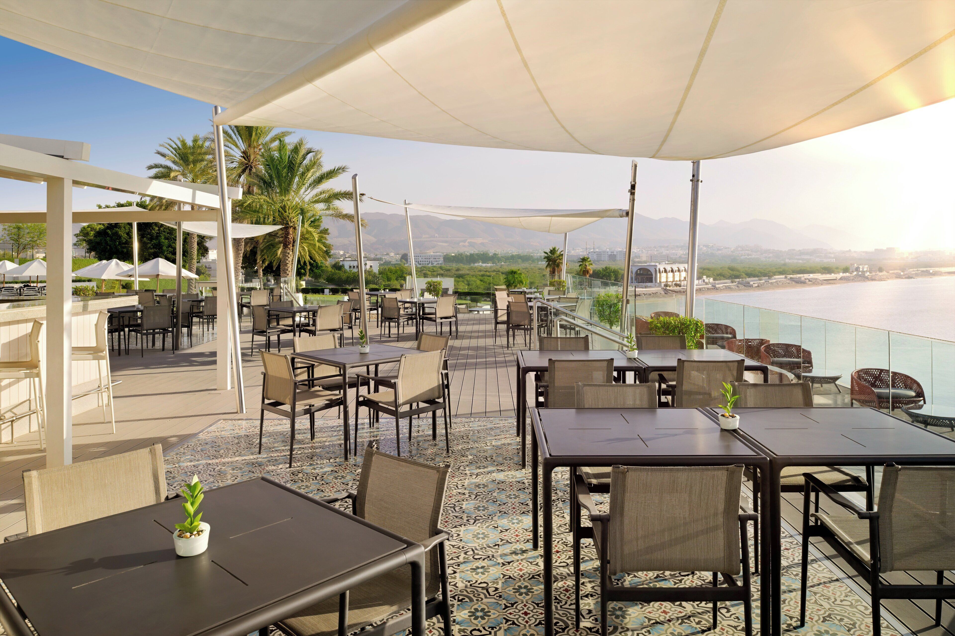 An outdoor dining area with tables and chairs, a view of the water, and palm trees.