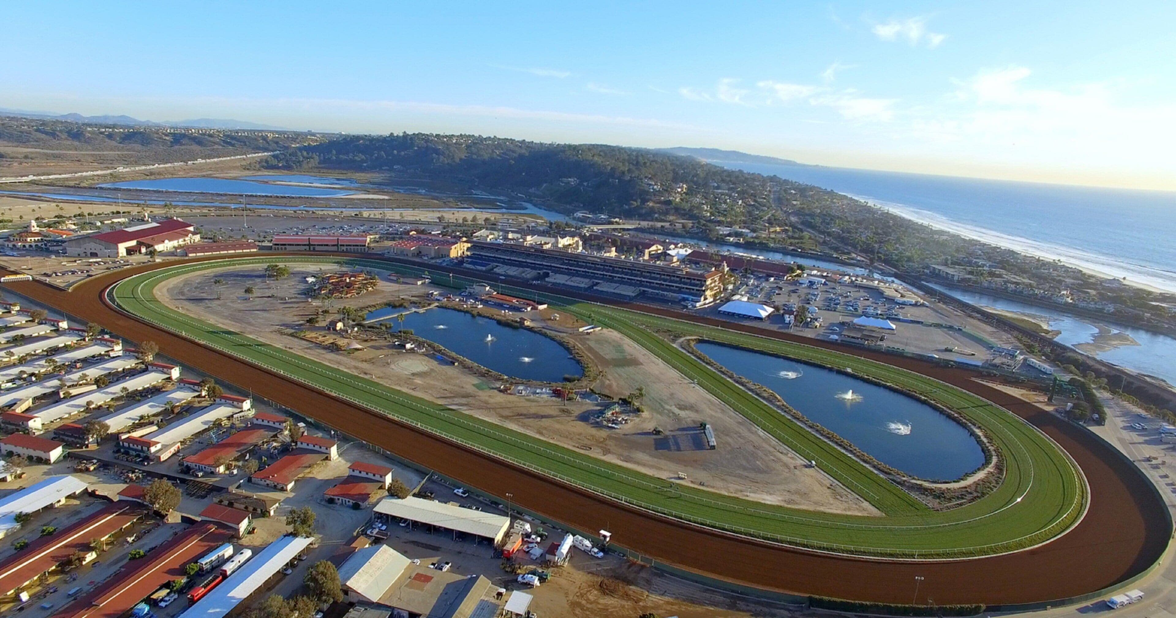 An aerial view of a racetrack with multiple lanes, spectator stands, and a coastal backdrop.