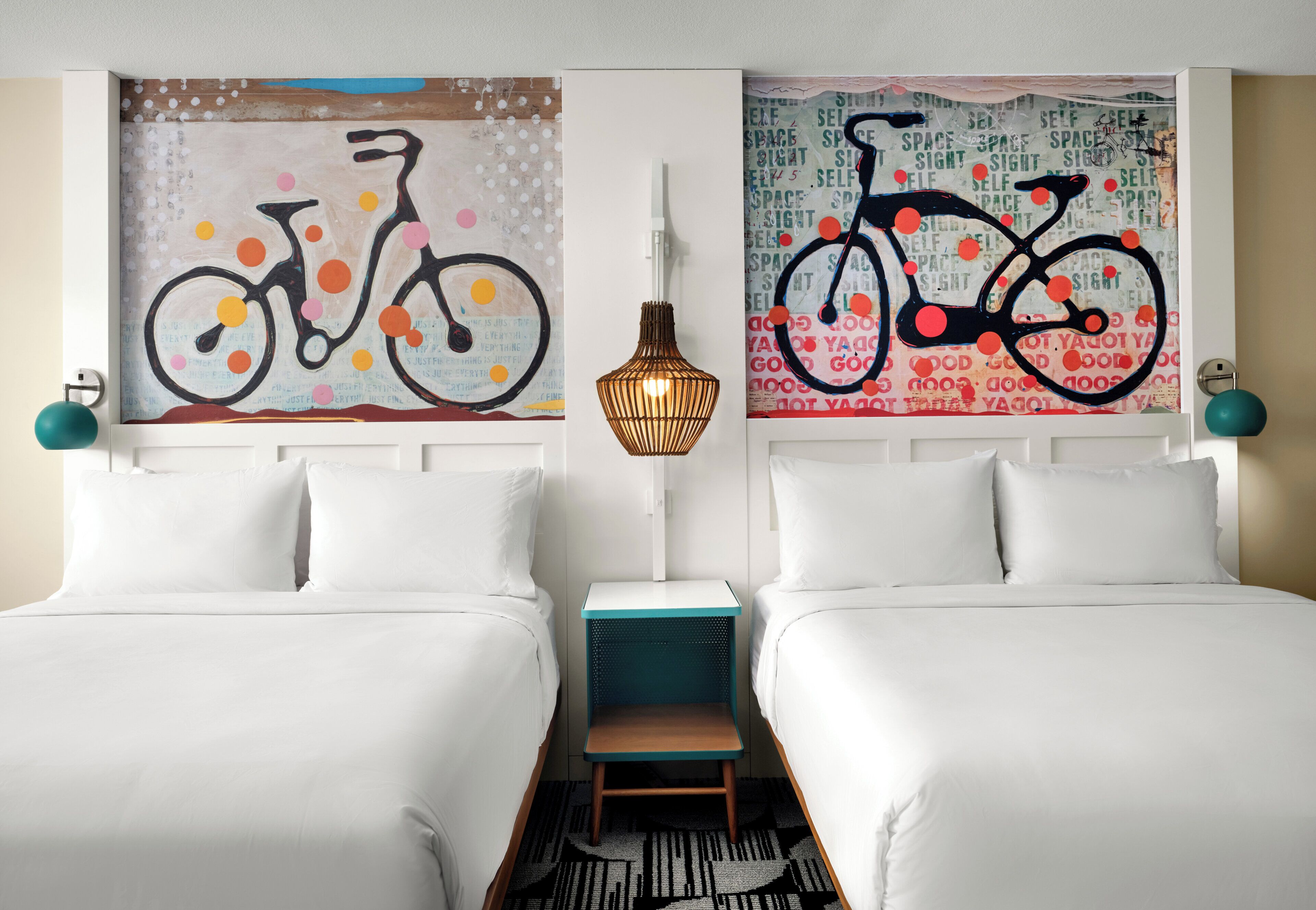 Une chambre d’hôtel avec deux lits, une lampe fixée au mur et des œuvres d’art représentant des bicyclettes.