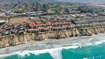 An aerial view of a coastal residential area with a beachfront hotel featuring multiple buildings with red roofs, situated on a cliff overlooking the ocean.