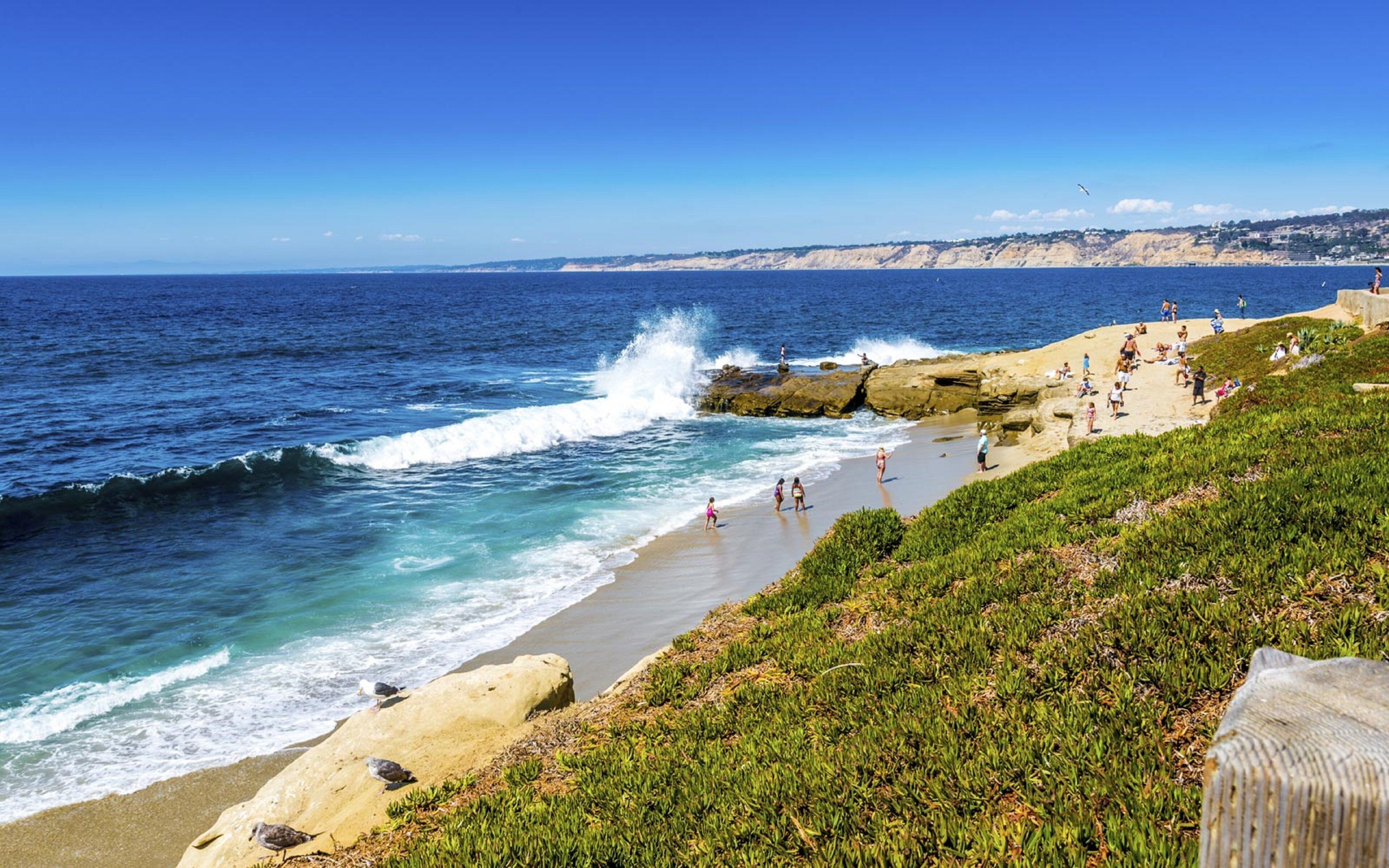 A coastal beach with waves crashing onto the shore, people walking along the beach, and a rocky cliff in the background.