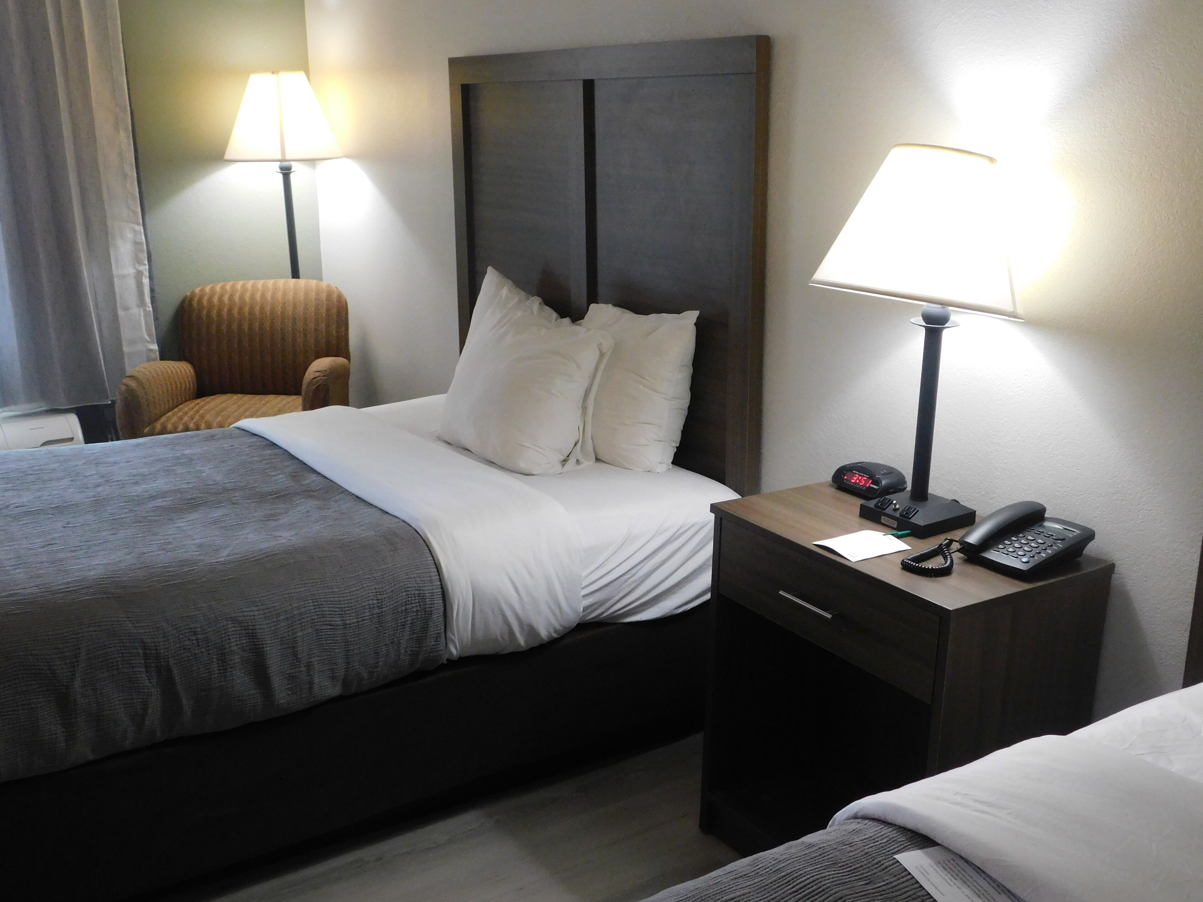 A hotel room with a bed, bedside table, lamp, chair, and a clock.