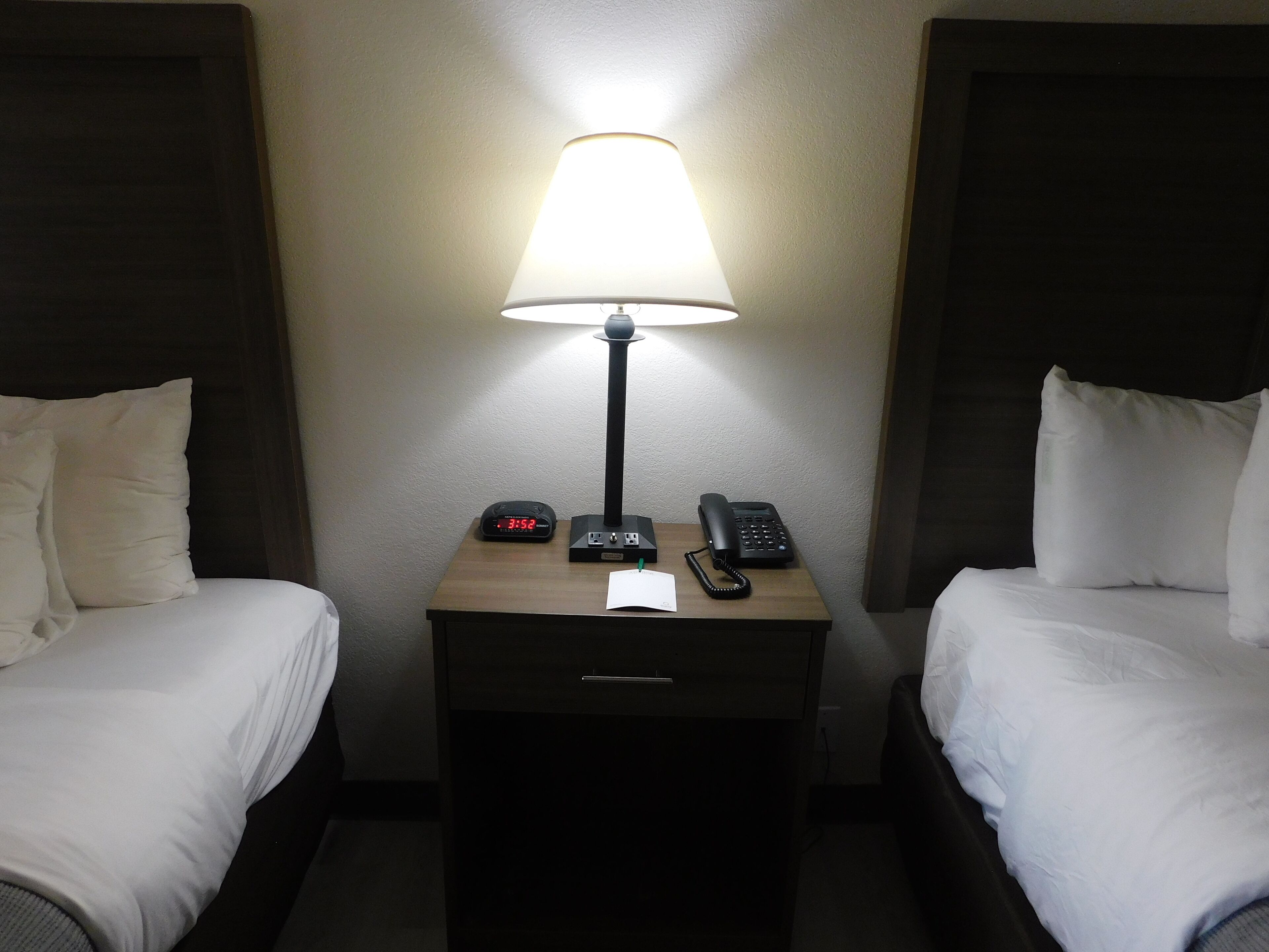 Habitación de hotel con dos camas, una mesita de noche con lámpara, un reloj y un teléfono.