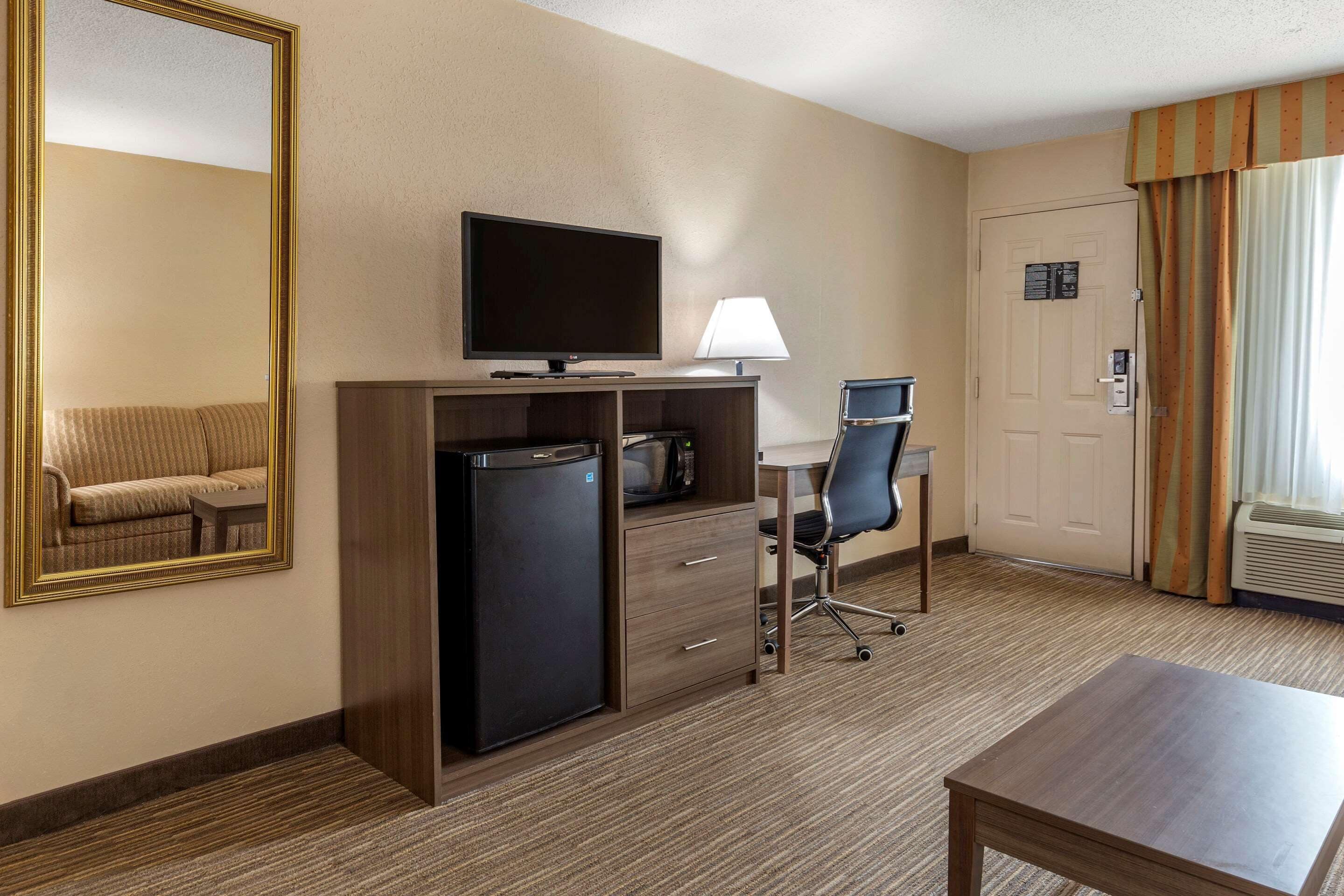 A hotel room with a large mirror, a flat-screen TV, a desk with a chair, a refrigerator, a lamp, and a bed.