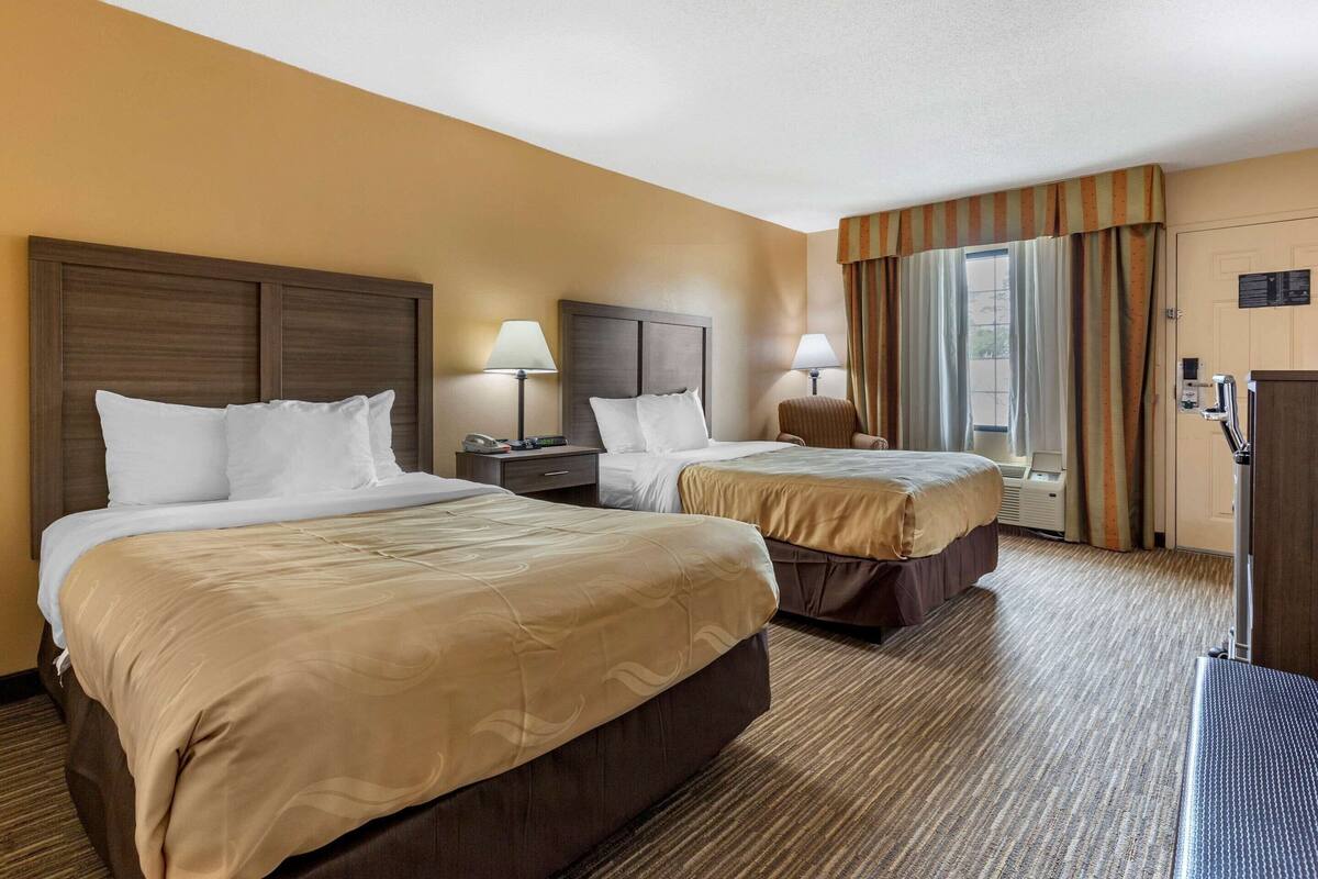 Habitación de hotel con dos camas, un escritorio, una silla, un ventanal con cortinas y una puerta.