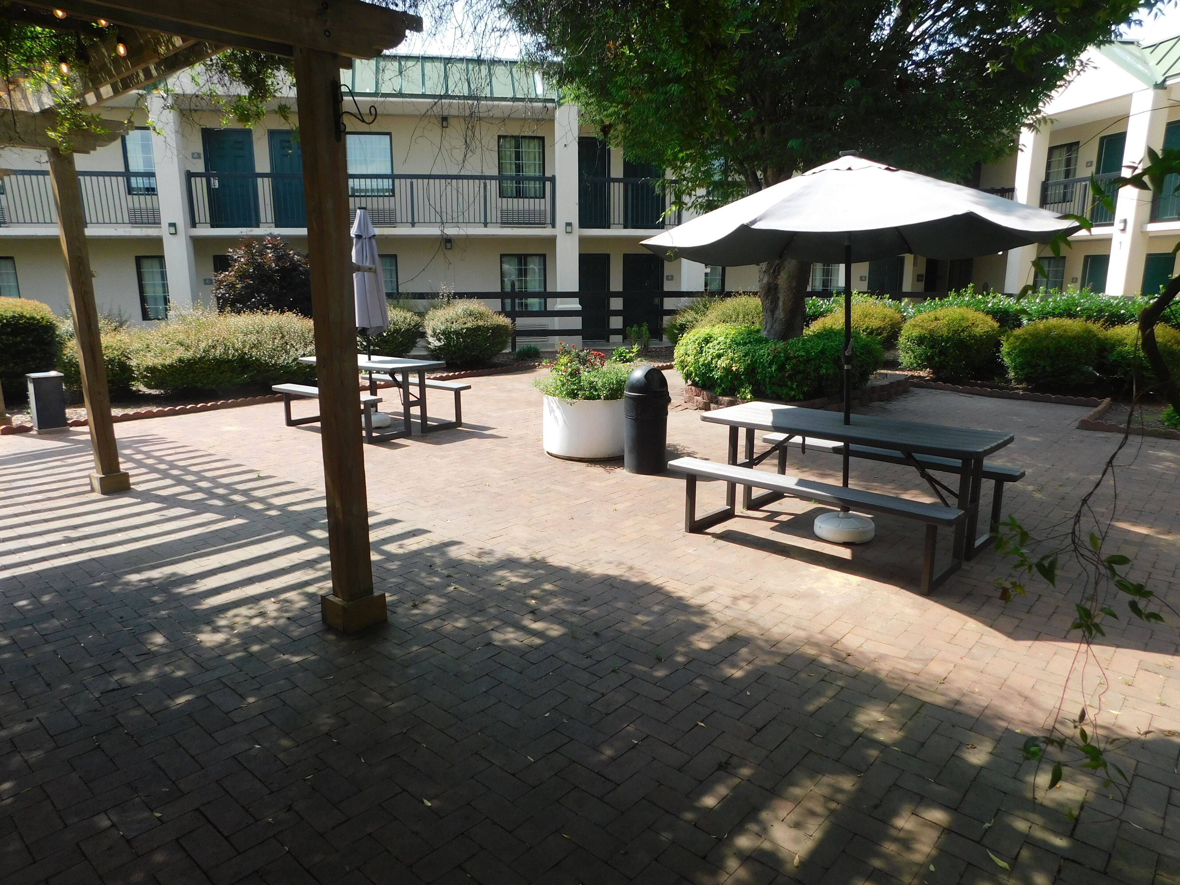An outdoor patio area with picnic tables and umbrellas, surrounded by greenery and a building in the background.