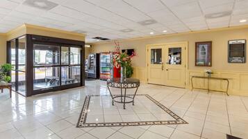 A hotel lobby with a tiled floor, a reception desk, a vending machine, and a glass door entrance.
