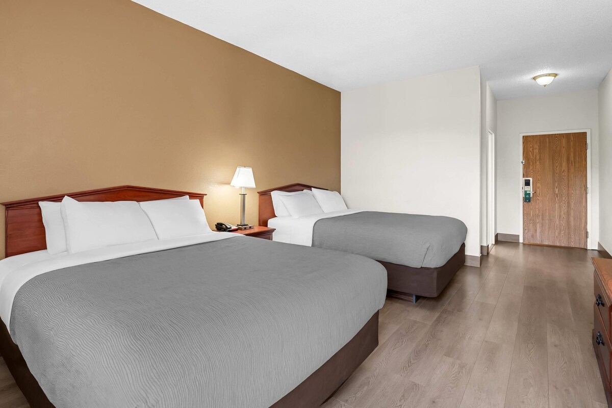 A hotel room with two beds, a bedside table with a lamp, a wooden door, and a hallway.