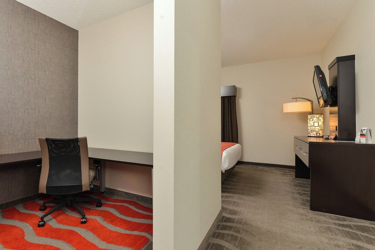 A hotel room with a desk, chair, bed, and a small table with a lamp.