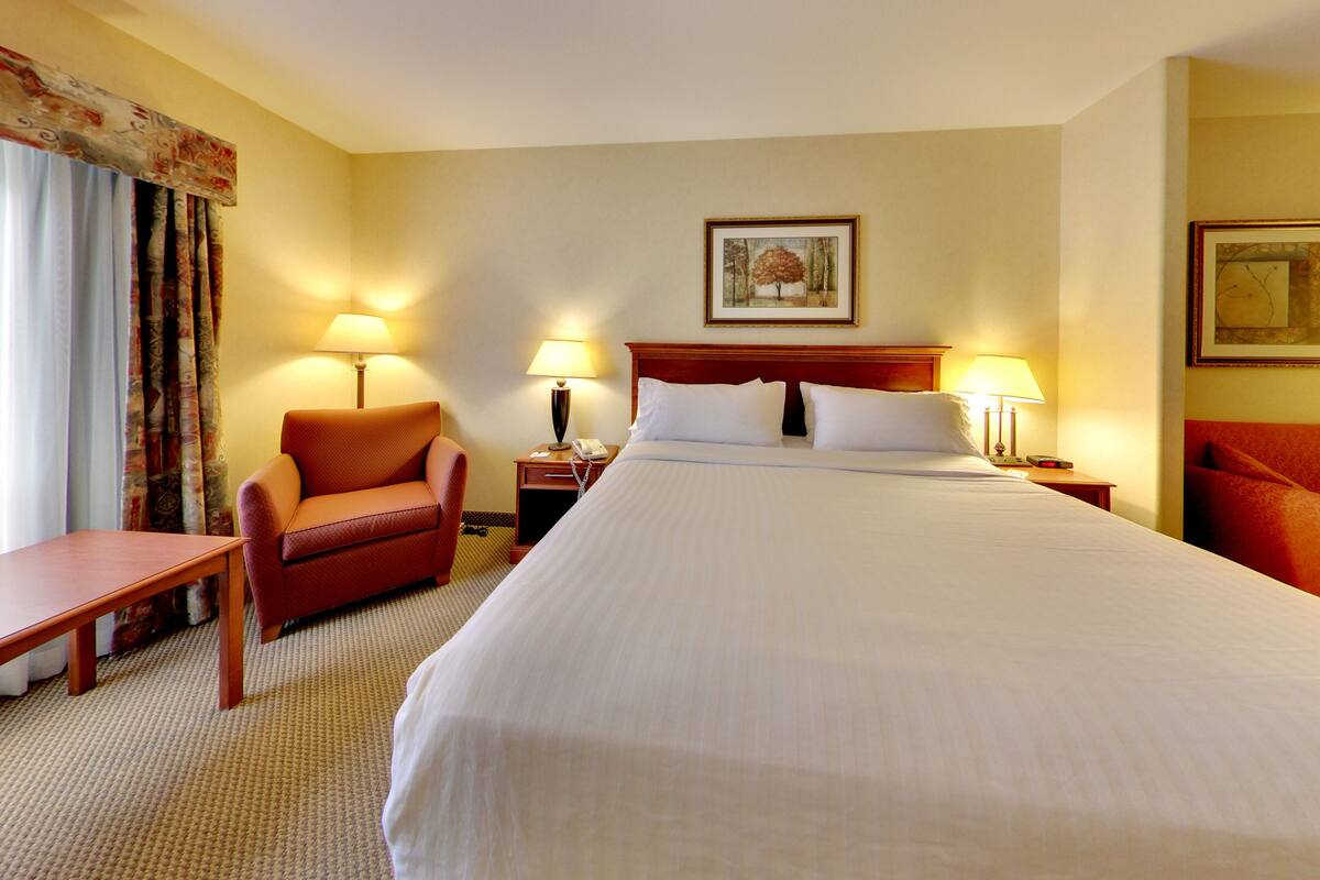 A hotel room with a large bed, a wooden chair, a small table, two bedside lamps, and a painting on the wall.