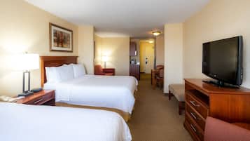 A hotel room with two beds, a TV, a desk with a lamp, a chair, and a small kitchenette.