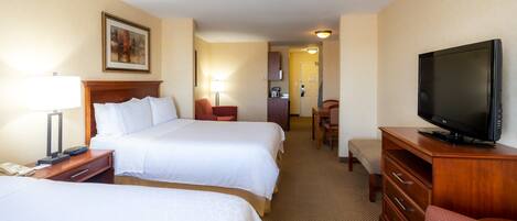 A hotel room with two beds, a TV, a desk with a lamp, a chair, and a small kitchenette.