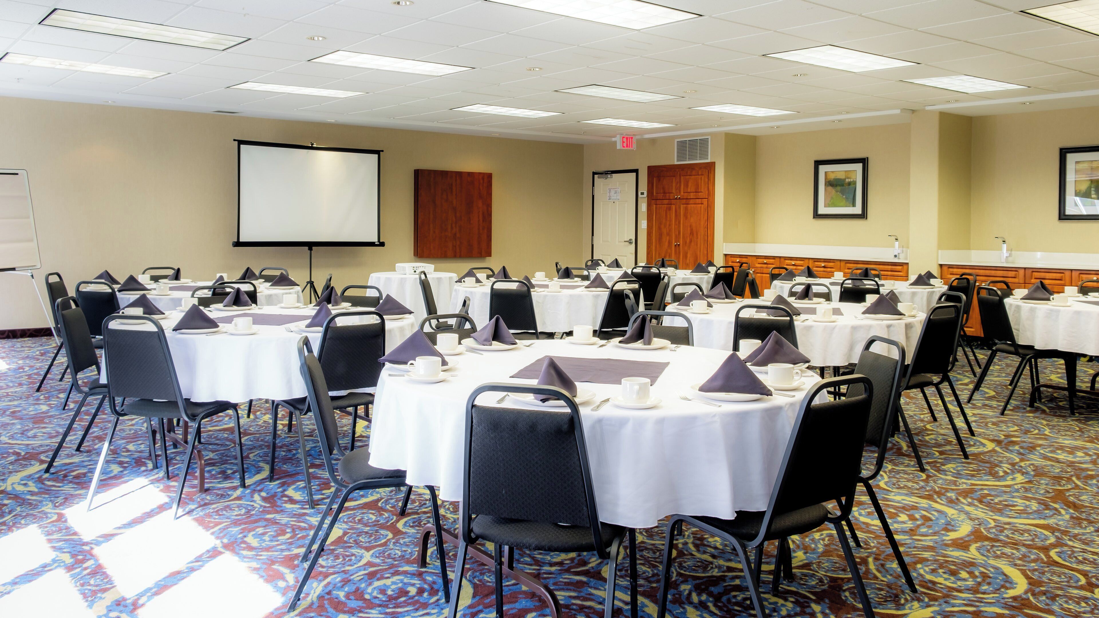 A conference room with multiple tables set for a meeting, equipped with nameplates, and a projector screen.