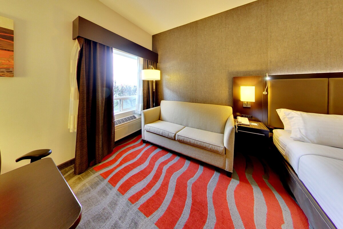 A hotel room with a sofa, a bed, a nightstand, a lamp, a window with curtains, and a patterned rug.