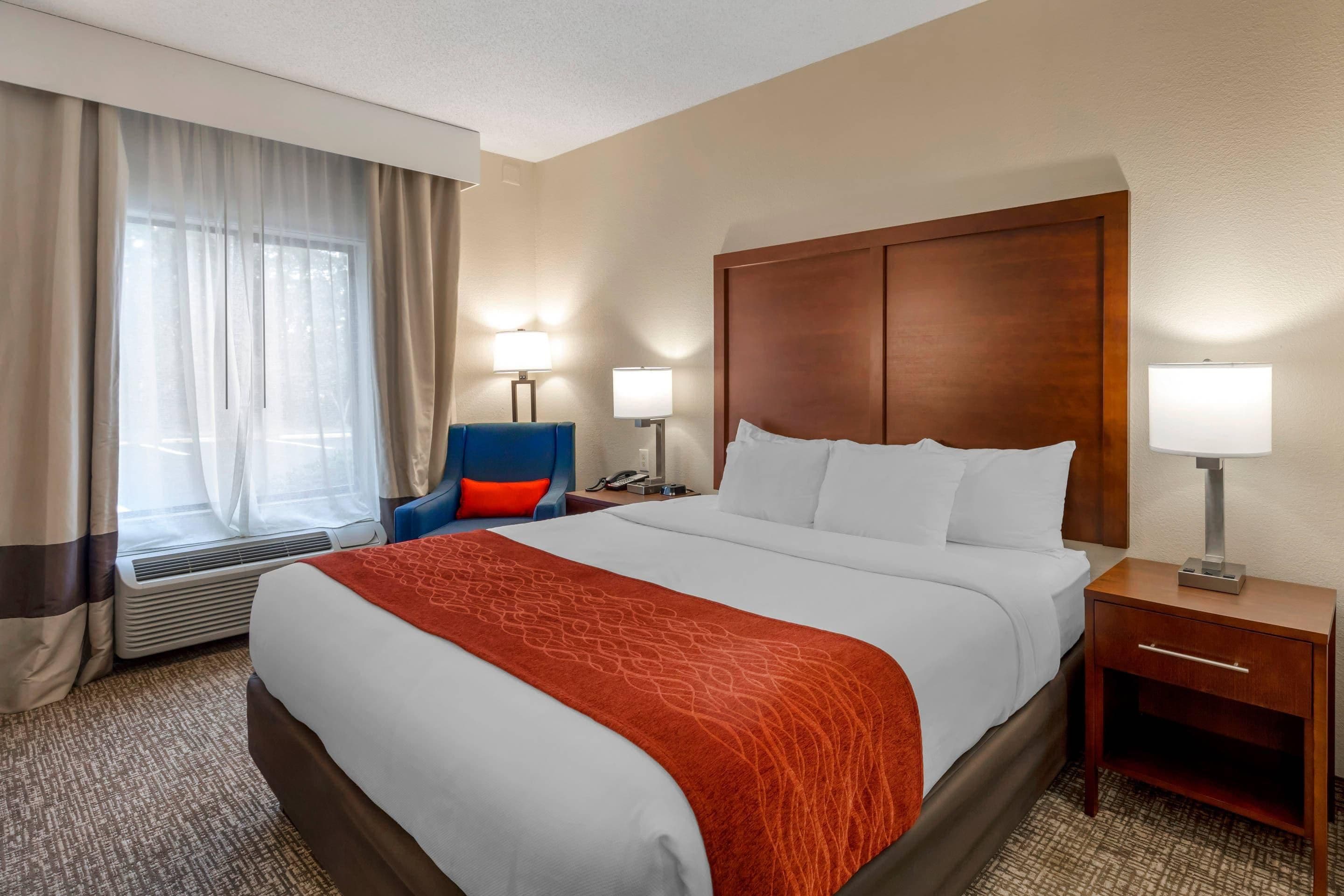Een hotelkamer met een groot bed, twee bedlampen, een houten hoofdeinde, een blauwe stoel, een kleine tafel en een raam met gordijnen.
