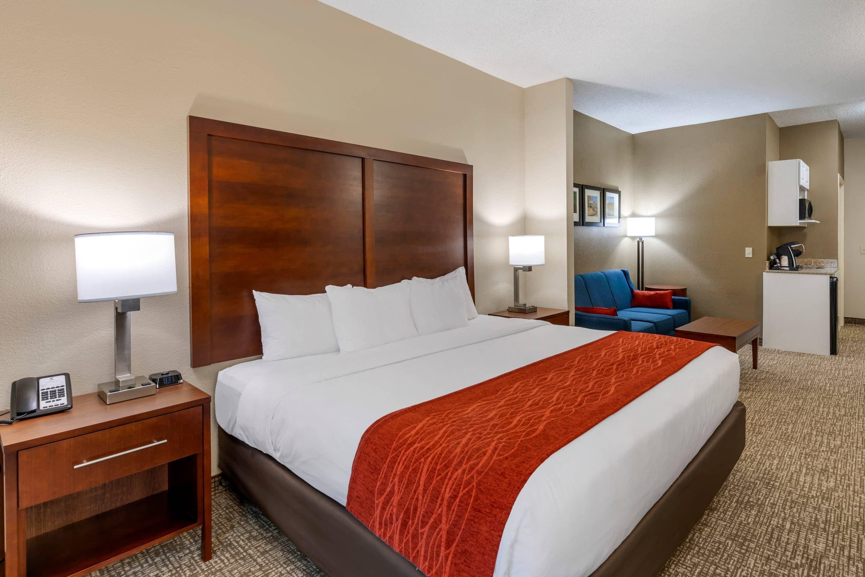 A hotel room with a large bed, a desk with a telephone, a chair, and a small kitchenette.