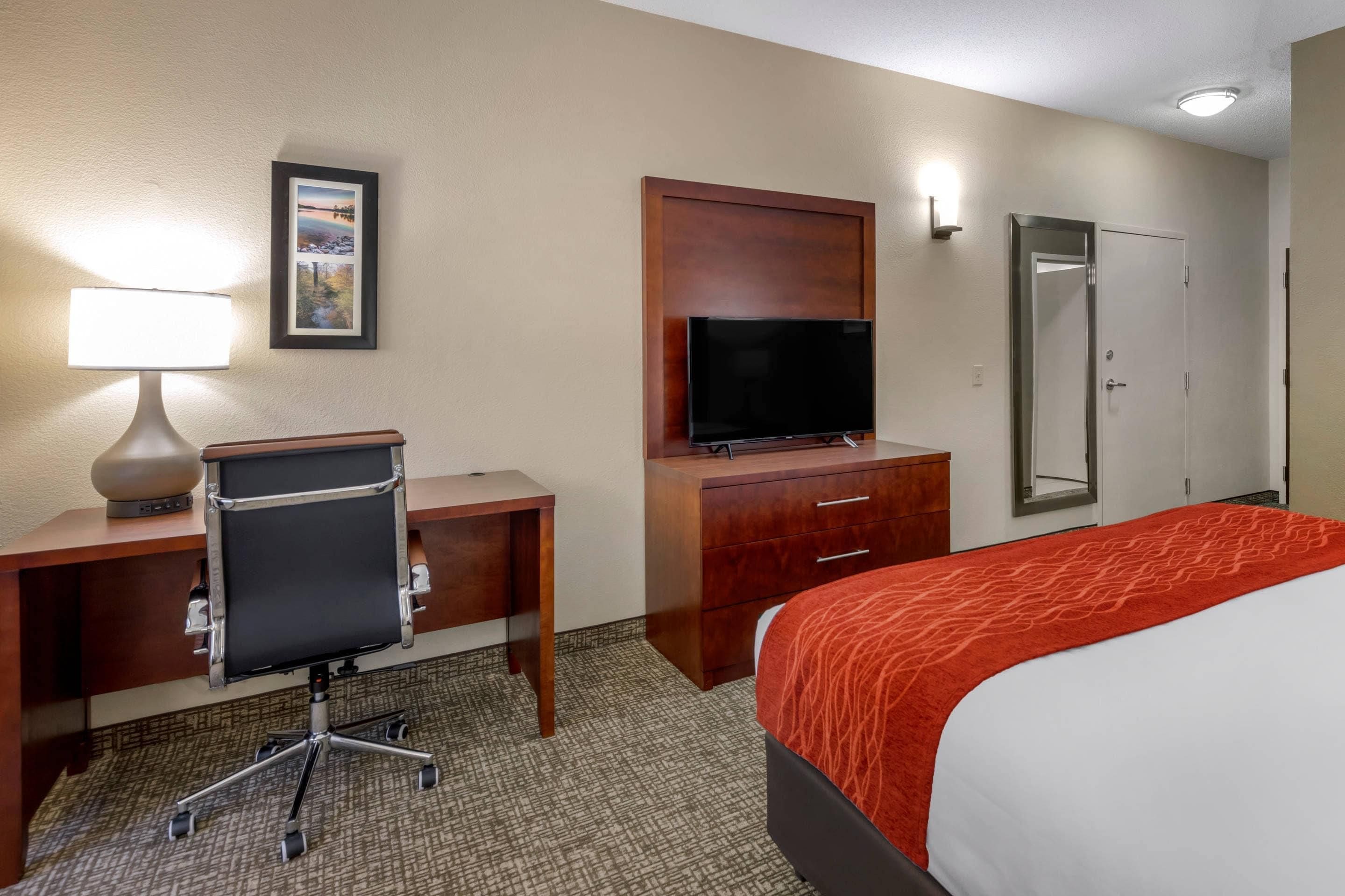 A hotel room with a desk, chair, television, wardrobe, and a bed with a red blanket.