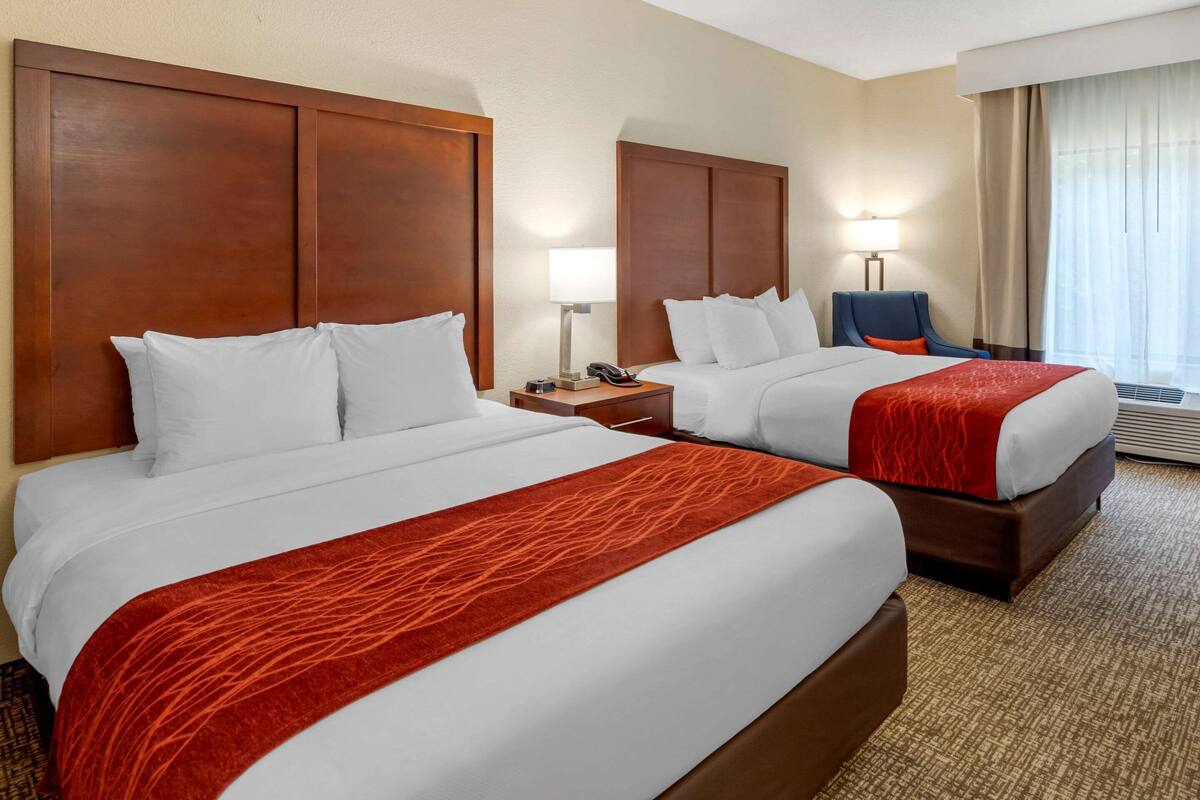 A hotel room with two beds, a nightstand with a lamp, a chair, and a large window with curtains.