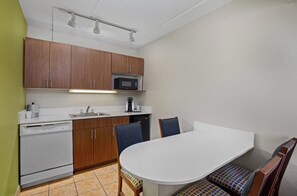 Cuina compacta amb armaris de fusta, microones, rentaplats i taula de dinar blanca amb cadires.