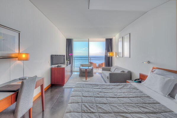 A modern hotel room with a large bed, a desk, a sofa, and a view of the ocean. - Vidamar Resort Madeira (Funchal)