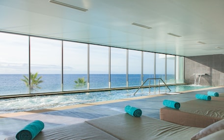 A modern indoor pool area with large windows overlooking the ocean, a small waterfall feature, and several lounge chairs with towels.