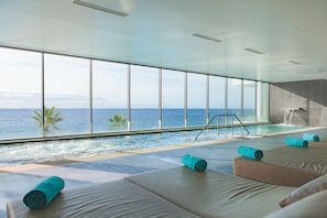 A modern indoor pool area with large windows overlooking the ocean, a small waterfall feature, and several lounge chairs with towels. - Vidamar Resort Madeira (Funchal)