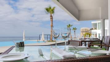 A dining table with a clear view of the ocean and palm trees, set with wine glasses and cutlery.