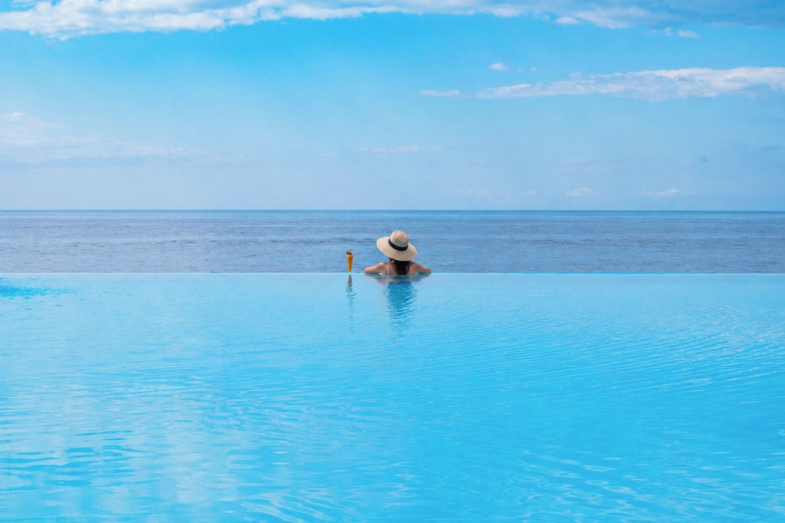 A clear blue pool with a person in the water, a hat on the edge, and a view of the ocean.