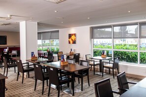 A conference room with multiple tables and chairs, large windows with a view of greenery and parked cars, and a flat-screen TV mounted on the wall. - Holiday Inn Express Glasgow Airport by IHG (Paisley)