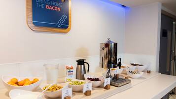 A breakfast buffet with labeled bowls of yogurt, butter, and other spreads, a coffee machine, and a sign that reads 'BRING HOME THE BACON'.