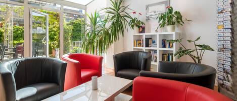 Un interior moderno con una pared de vidrio, sillas negras y rojas, una mesa blanca y una estantería con libros y plantas.