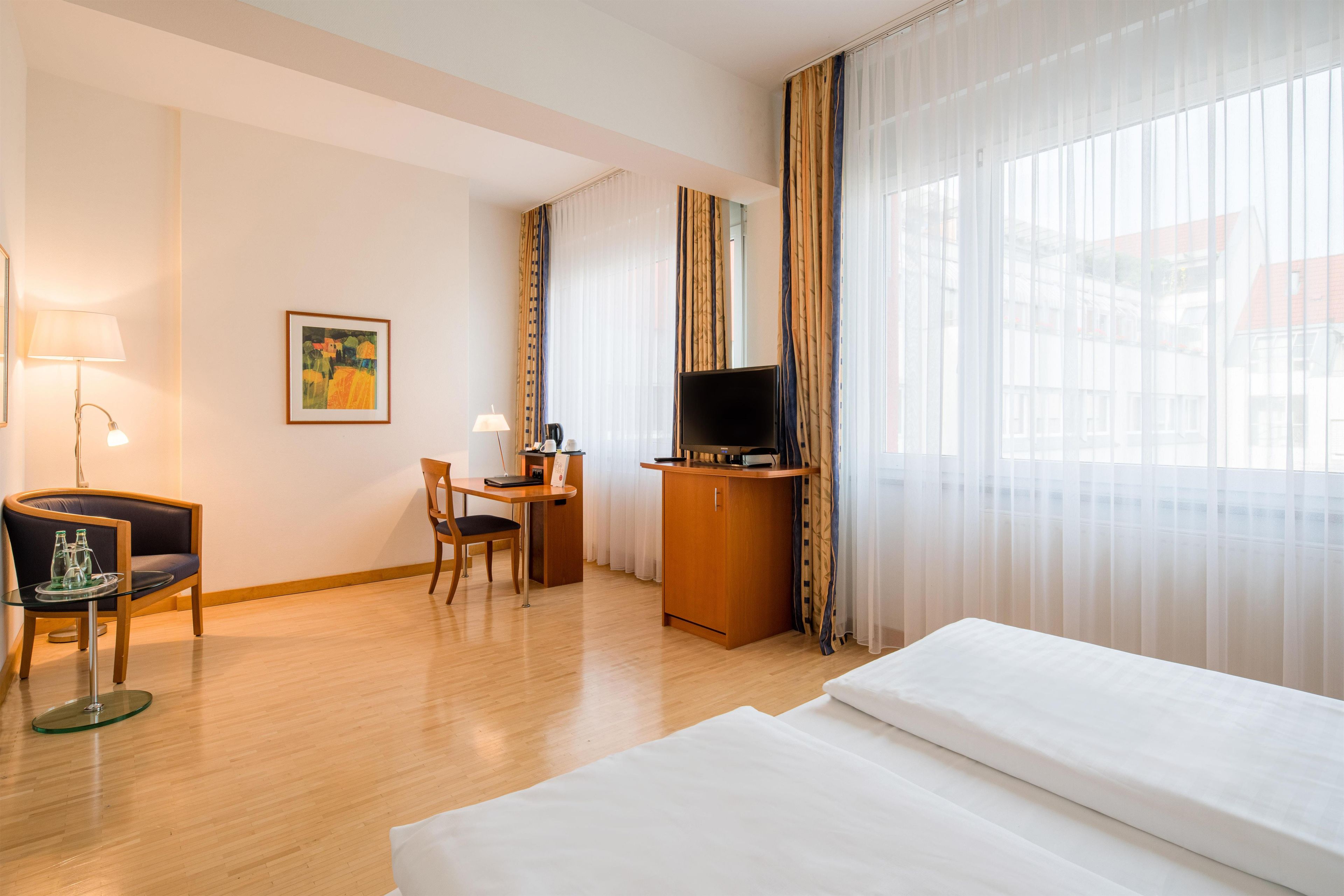 A hotel room with a bed, a desk with a TV, a chair, a lamp, and a painting on the wall.