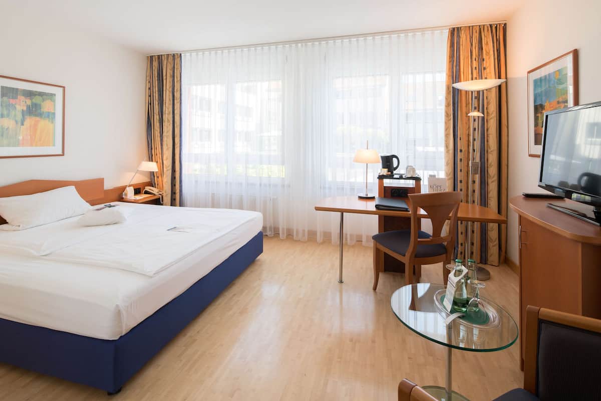 A hotel room with a bed, desk, chair, TV, and a glass table with a bottle.