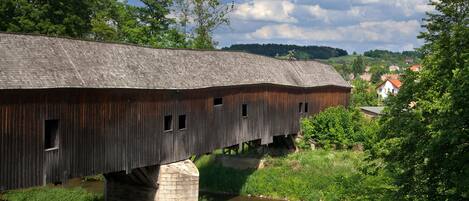 A wooden covered bridge over a river with a stone foundation, surrounded by greenery and houses in the background.